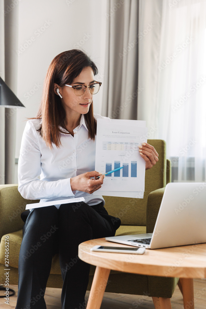 Concentrated beautiful young business woman in formal wear clothes indoors at home talking using laptop computer with earphones showing graphics. Concentrated beautiful young business woman in formal wear clothes indoors at home talking using laptop computer with earphones showing graphics.
