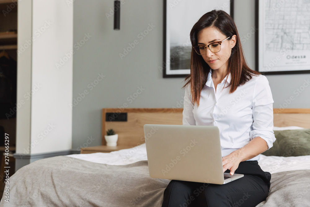 Portrait of concentrated adult businesswoman typing on laptop while sitting on bed in apartment Portrait of concentrated adult businesswoman typing on laptop while sitting on bed in apartment