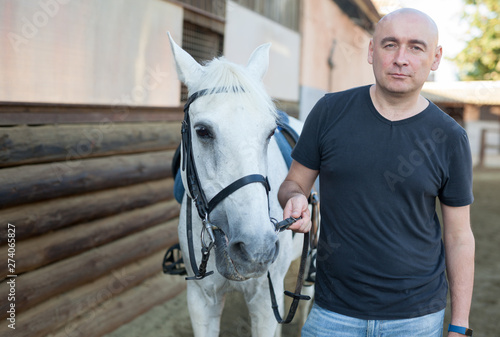 Mature smiling man farmer standing with white horse at stable
