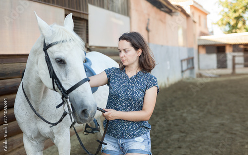 Mature smiling woman farmer standing with white horse at stable