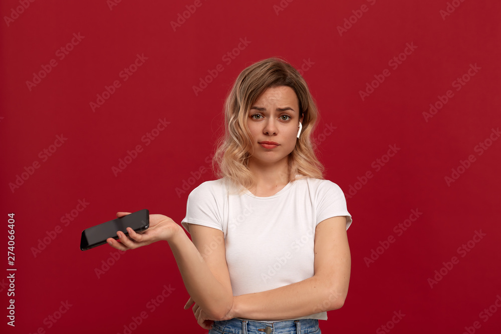 Portrait of a girl with curly blond hair in a white t-shirt on a red background. Unhappy model in wireless headset holds mobile phone. Portrait of a girl with curly blond hair in a white t-shirt on a red background. Unhappy model in wireless headset holds mobile phone.