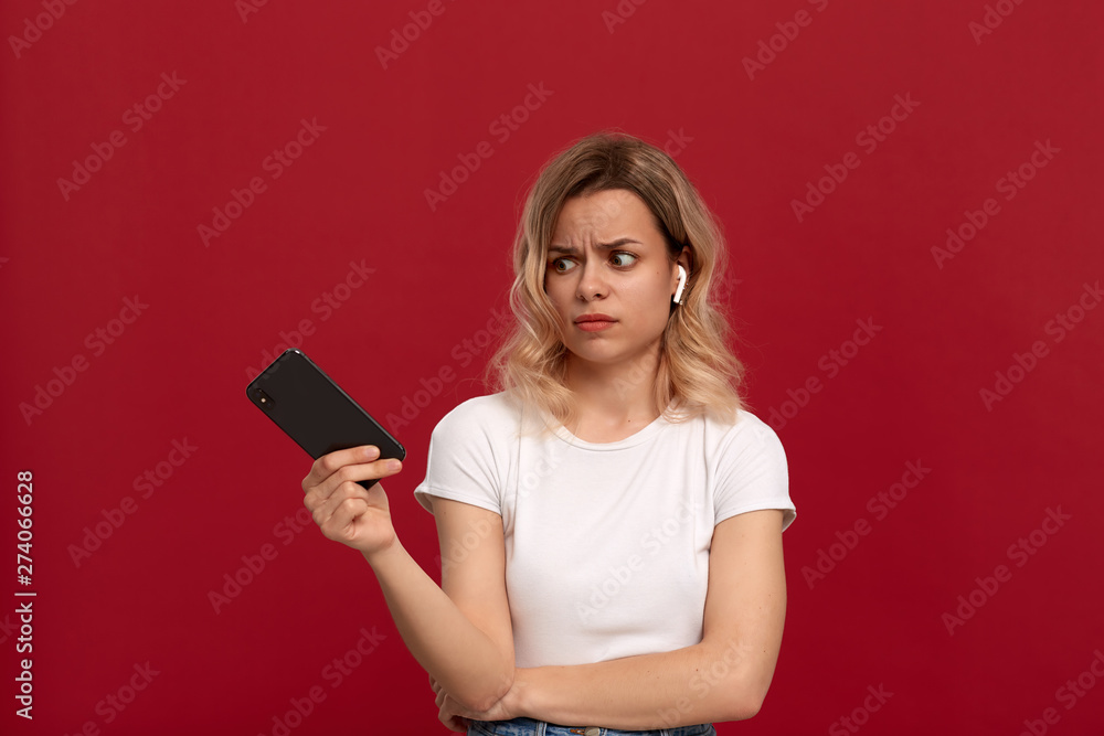 Portrait of a girl with curly blond hair in a white t-shirt on a red background. Model with dissatisfied look in wireless headset looks at the screen of a mobile phone. Portrait of a girl with curly blond hair in a white t-shirt on a red background. Model with dissatisfied look in wireless headset looks at the screen of a mobile phone.