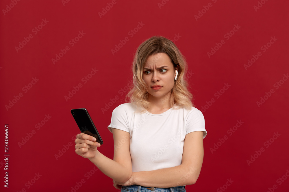 Naklejka premium Portrait of a girl with curly blond hair in a white t-shirt on a red background. Model with dissatisfied look in wireless headset looks at the screen of a mobile phone.