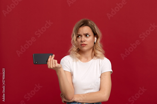 Portrait of a girl with curly blond hair in a white t-shirt on a red background. Model with dissatisfied look in wireless headset holds mobile phone.