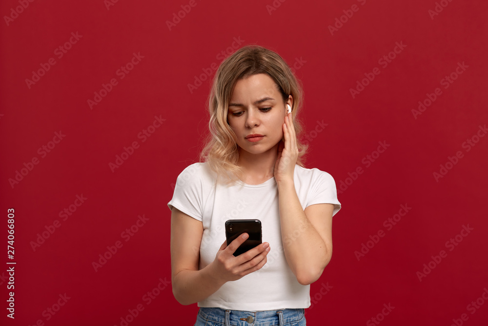 Portrait of a girl with curly blond hair in a white t-shirt on a red background. Model in wireless headset looks at the screen of a mobile phone. Portrait of a girl with curly blond hair in a white t-shirt on a red background. Model in wireless headset looks at the screen of a mobile phone.