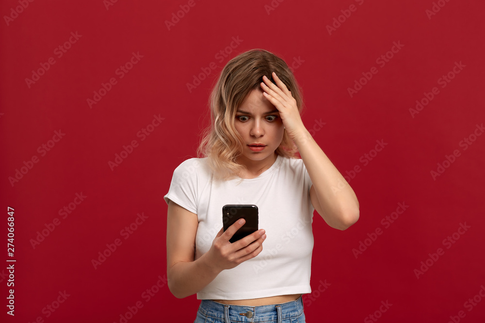 Portrait of a girl with curly blond hair in a white t-shirt on a red background. Model with dissatisfied look in wireless headset looks at the screen of a mobile phone. Portrait of a girl with curly blond hair in a white t-shirt on a red background. Model with dissatisfied look in wireless headset looks at the screen of a mobile phone.
