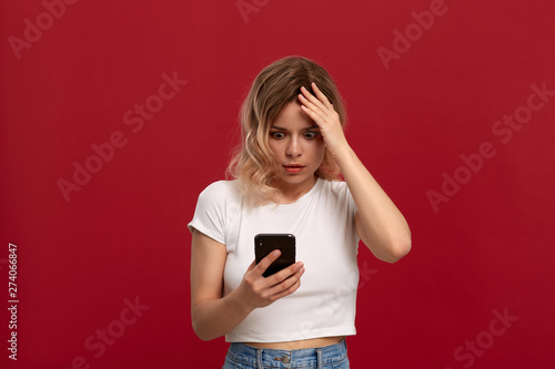 Portrait of a girl with curly blond hair in a white t-shirt on a red background. Model with dissatisfied look in wireless headset looks at the screen of a mobile phone.