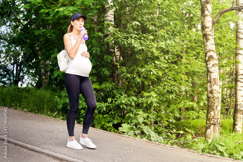 A pregnant young woman in a tracksuit and a baseball cap walks in the Park among the greenery and trees during sports training