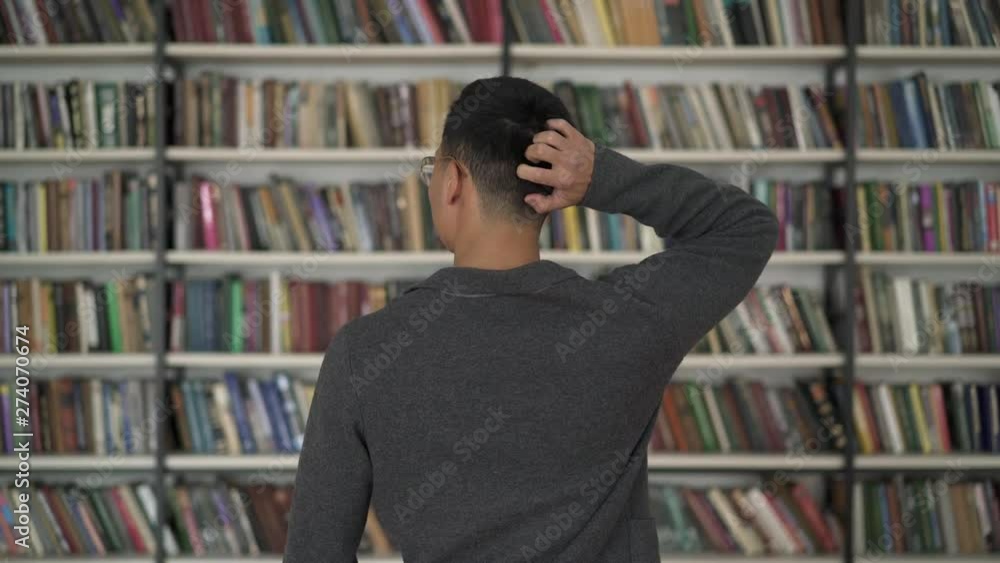 Gimbal middle shot of back of colored man choosing a book from bookshelf scratching his head. Asian man searching for a book on bookshelves in library scratching his head