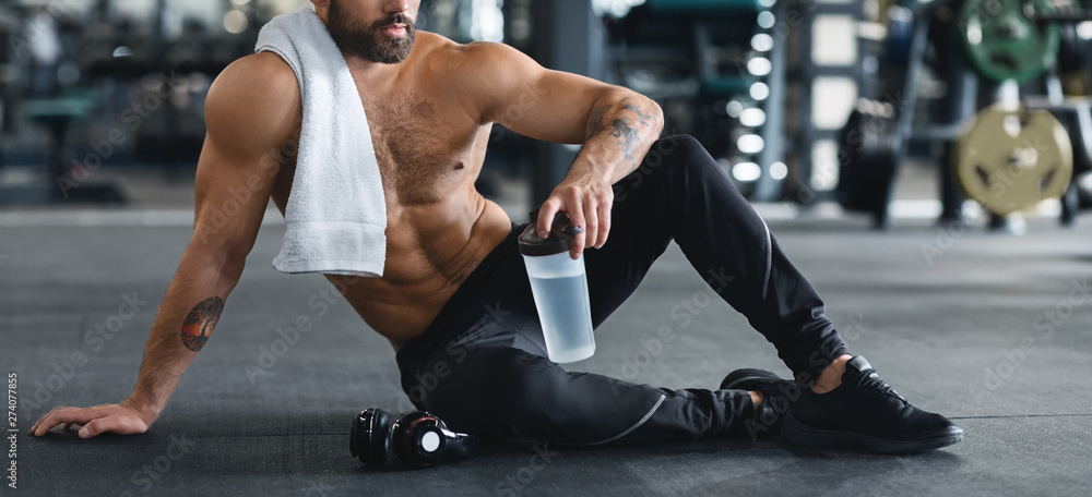 Naked man holding water bottle, sitting on floor at gym