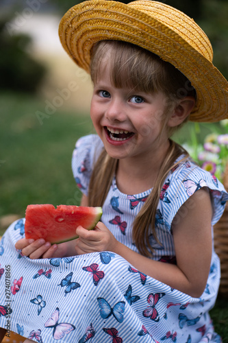 little beautiful girl of 4 years old in a dress and hat is eating a watermelon in the summer in the park at a picnic and smiling