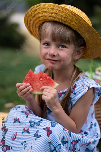 little beautiful girl of 4 years old in a dress and hat is eating a watermelon in the summer in the park at a picnic and smiling