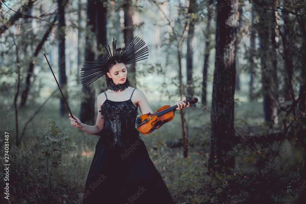 elegant woman in witch costume standing on forest background, looking at violin