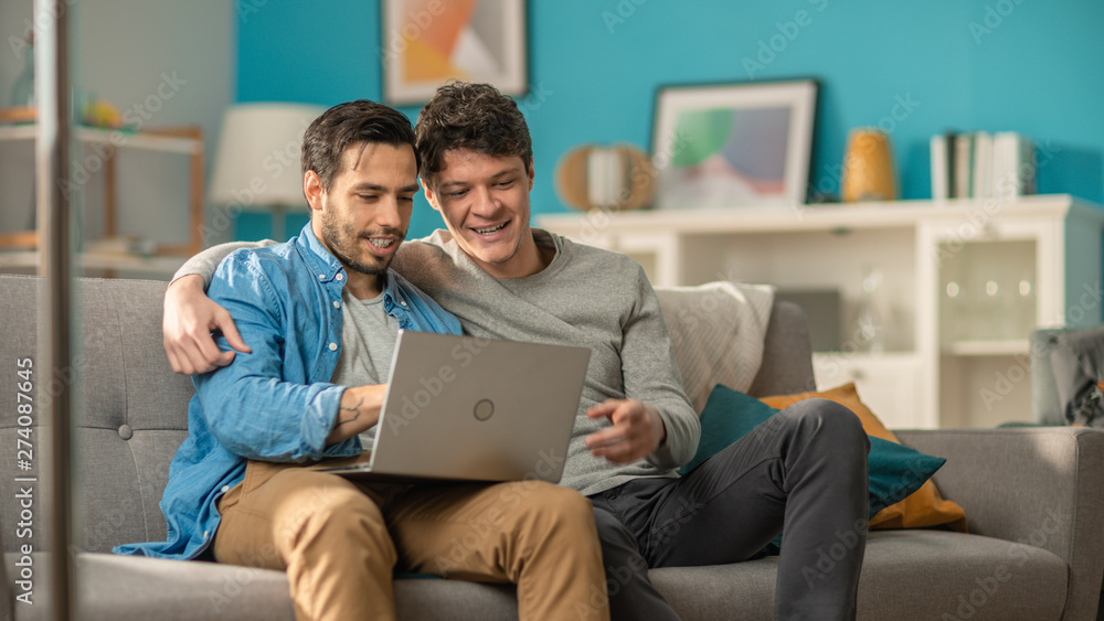 Adorable Male Gay Couple Spend Time at Home. They Sit on a Sofa and Use the Laptop. They Browse Online. Partner Puts His Hand Around His Lover. Room Has Modern Interior.