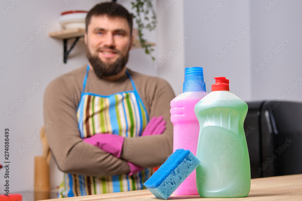 Presentation of different detergents with washcloth on the foreground, caucasian bearded man is standing in the background with his arms crossed Presentation of different detergents with washcloth on the foreground, caucasian bearded man is standing in the background with his arms crossed