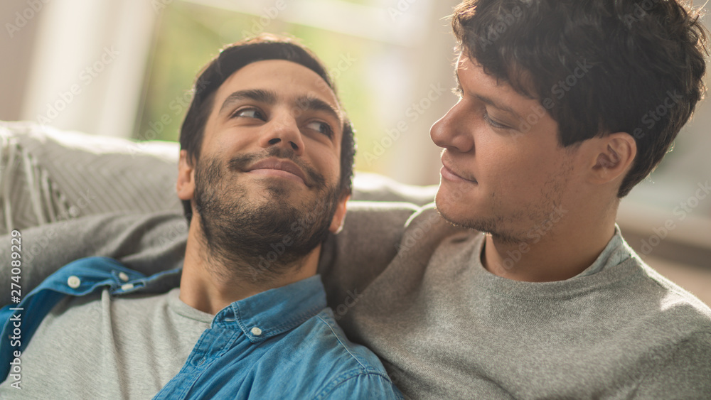 Sweet Male Queer Couple Spend Time at Home. They are Lying Down on a Sofa and Looking at Each Other Eyes. Partner's Hand is Around His Lover. They are Smiling. Room Has Modern Interior. Sweet Male Queer Couple Spend Time at Home. They are Lying Down on a Sofa and Looking at Each Other Eyes. Partner's Hand is Around His Lover. They are Smiling. Room Has Modern Interior.