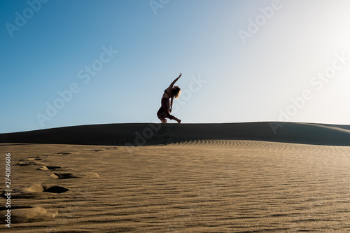 young woman with long skirt dancing in the distance in evocative and confident way on top of desert dune with clear blue sky