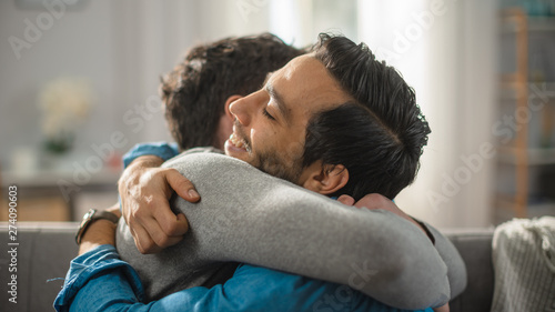 Cute Attractive Male Gay Couple Sit Together on a Sofa at Home. Boyfriends are Hugging and Embracing Each Other. They are Happy and Smiling. They are Casually Dressed and Room Has Modern Interior.