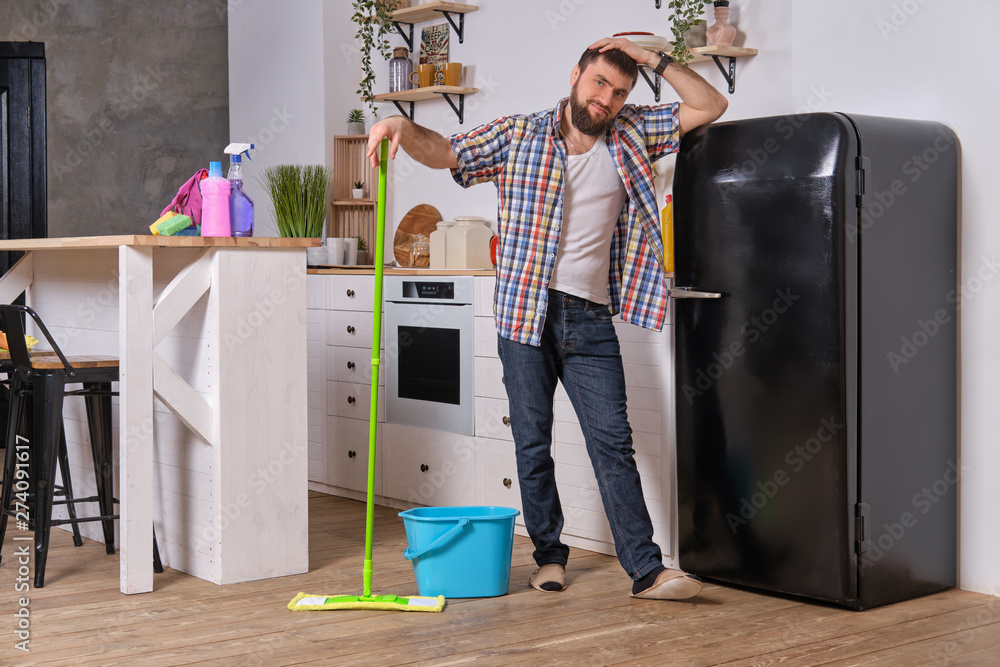 Husband housekeeper cleans the kitchen. Young happy man with green mop washes the floor Husband housekeeper cleans the kitchen. Young happy man with green mop washes the floor
