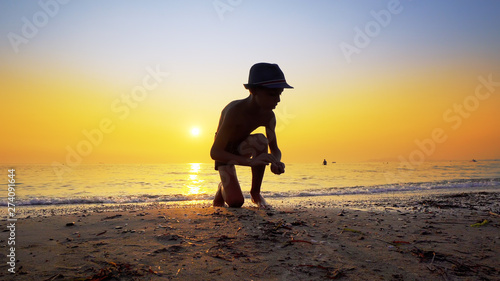 Young boy with hat collecting peebles on beach and throw stone skipping game on sea sunset water surface, small flattened rock bouncing off water surface across body of water many times