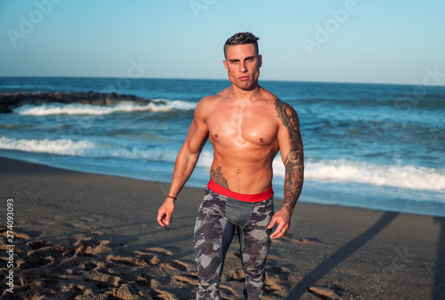 Tattooed bodybuilder sexy male coach at the beach. He stands and shows his muscles © Igor Kardasov