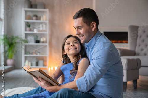 Horizontal shot of smiling father that reading book with his cute little daughter at home. Little cheerful girl look at her dad