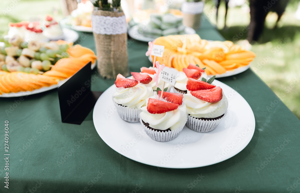 Details of a festival catering: fruits, cupcakes, macaroons and burgers. Sweet table at the feast. Delicious candy bar at the wedding ceremony close up. Catering service plate. Wedding party concept