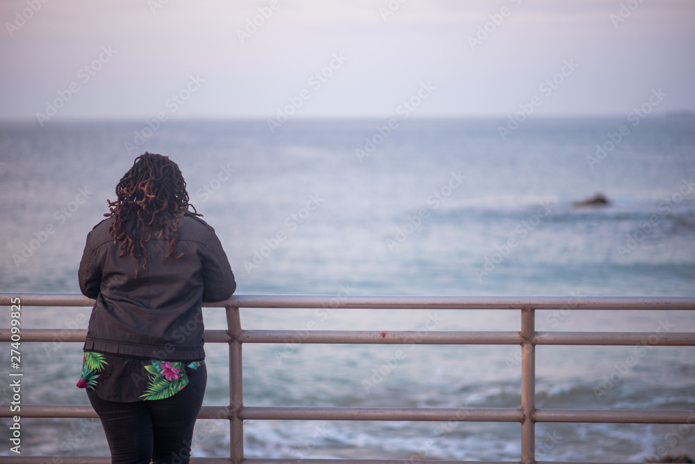 Woman with dreadlocks standing and watching the ocean. Shot from behind