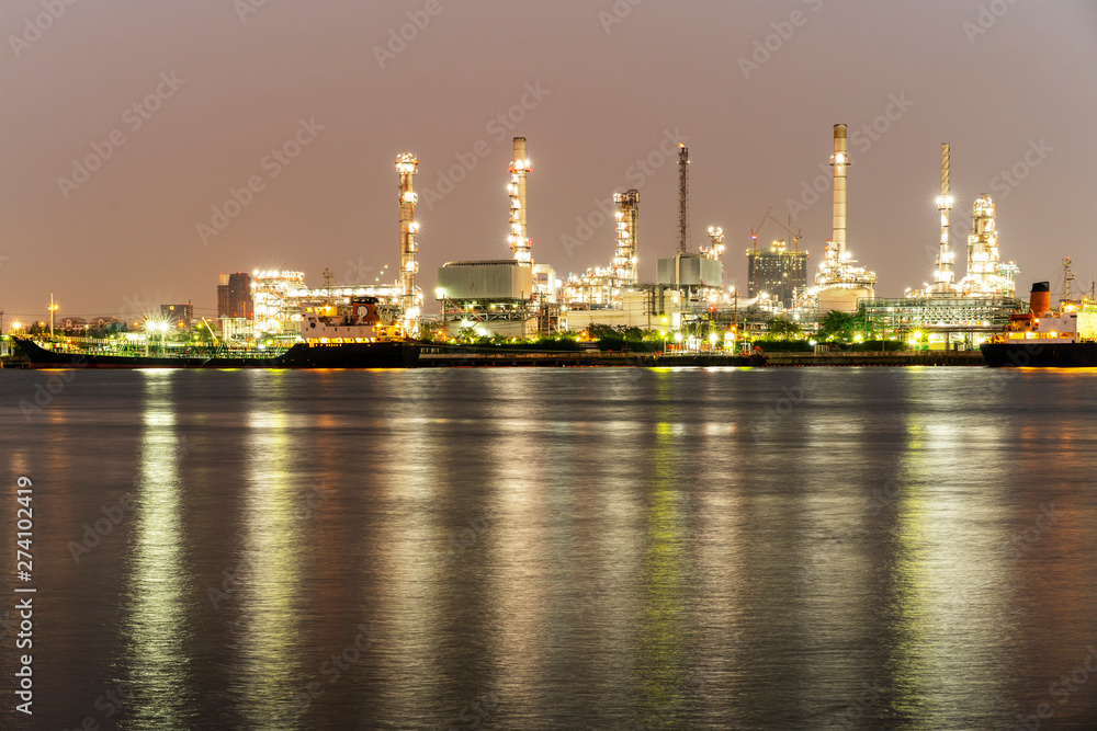 Night photo crude oil refinery plant and many chimney with petrochemical tanker or cargo ship at coast of the river under colorful golden bright lights at thailand