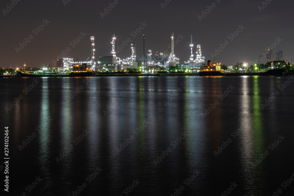 Night photo crude oil refinery plant and many chimney with petrochemical tanker or cargo ship at coast of the river with colorful bright light from lamp reflect on water at thailand