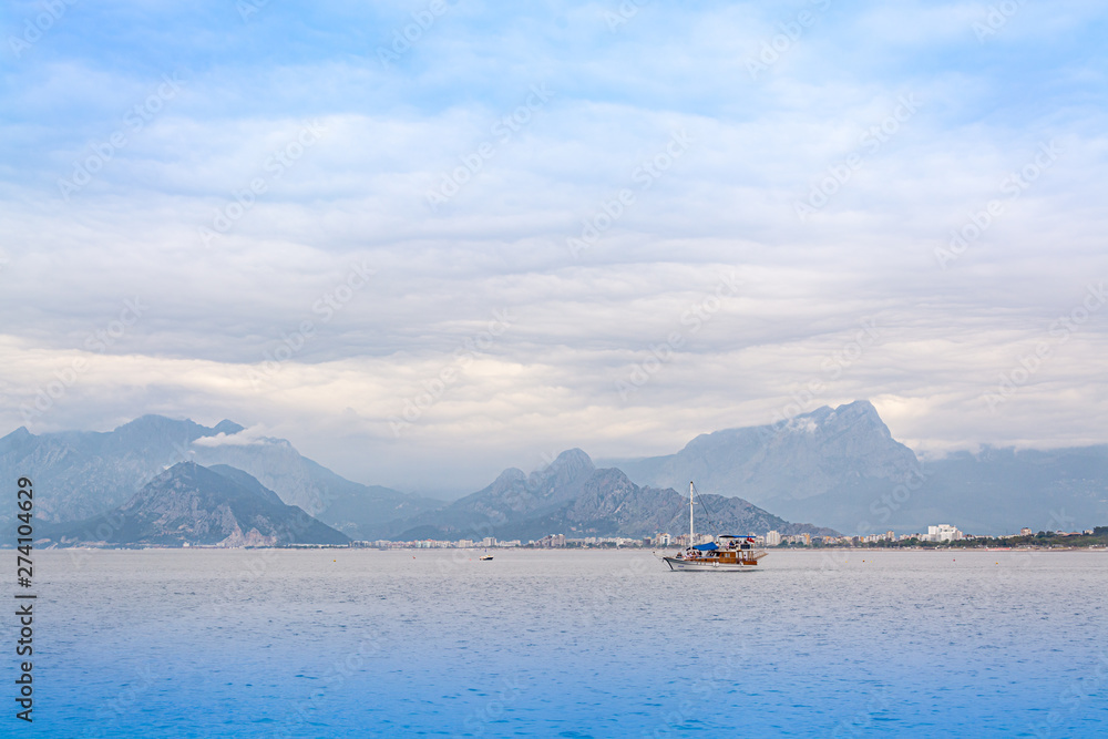 One tourist boat floats in the ocean or the sea against the backdrop of mountains. Cloudy sky. High mountains run into the clouds. At the foot of the mountains you can see a lot of houses.