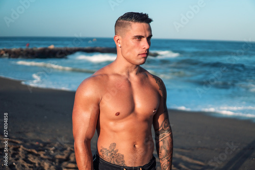 Tattooed bodybuilder sexy male coach at the beach. He stands and shows his muscles