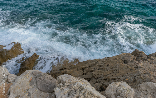 Summer cliff next to the Mediterranean Sea, strong waves break with the rocks and leave blue and turquoise colors along with the foam of the sea.