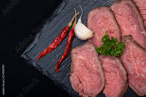 Sliced Grass Fed Corn Roast Beef garnished with Garlic, Fresh Curly Parsley and dried Red Chile De Arbol Peppers on natural black stone background.