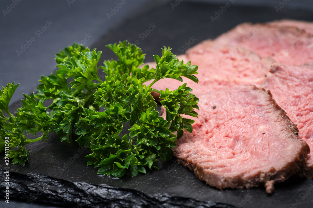 Sliced Grass Fed Juicy Corn Roast Beef garnished with Fresh Curly Parsley on black natural stone background. Sliced Grass Fed Juicy Corn Roast Beef garnished with Fresh Curly Parsley on black natural stone background.