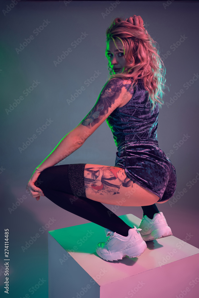 Young fitness blonde woman wearing blue velour booty shorts posing on camera,  making squats on top of big cube foto de Stock | Adobe Stock