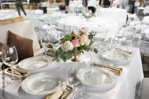 Wedding table setting decorated with fresh flowers in a brass vase. Wedding floristry. Banquet table for guests outdoors with a view of green nature. Bouquet with roses, eustoma and eucalyptus leaves