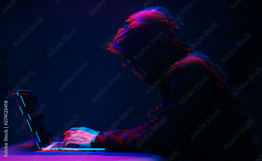 Hacker in the hood working with laptop typing text in dark room. Image with glitch effect