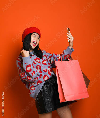 Asian woman in a stylish beret and sweater, with a colorful shopping bag makes a hard-working selfie on the phone after shopping