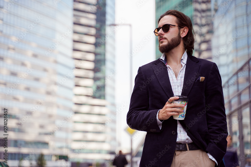 Photo of brunet with beard and earring in ear looking to side with glass in hand in city against background of tall buildings Photo of brunet with beard and earring in ear looking to side with glass in hand in city against background of tall buildings