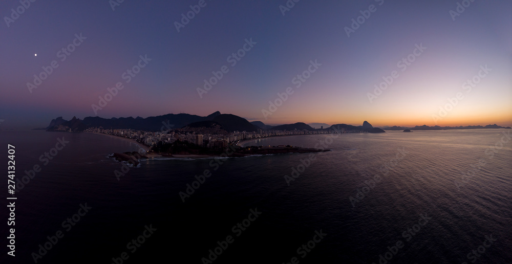 Aerial wide panorama of Rio de Janeiro with Arpoador cliff and wider cityscape and skyline in the background against a colourful sunrise sky with the moon visible above the city on the left Aerial wide panorama of Rio de Janeiro with Arpoador cliff and wider cityscape and skyline in the background against a colourful sunrise sky with the moon visible above the city on the left