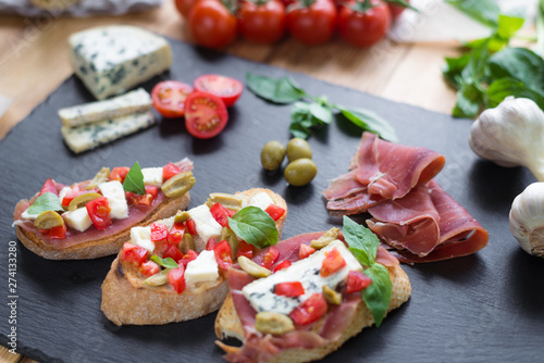 Traditional Italian bruschetta with blue cheese, feta, tomatoes, basil leaves, jamon on black stone background.