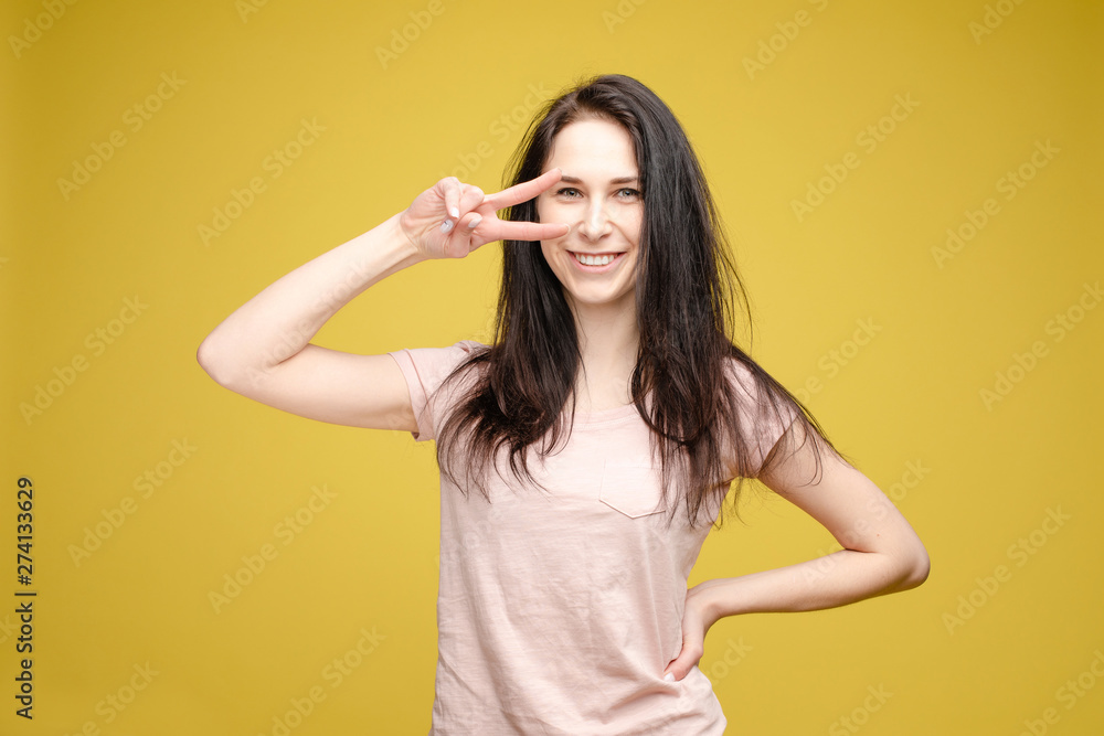 Studio portrait of charming brunette young woman in white top and green skirt holding peace sign near her eye. She is smiling at camera. Isolate on orange. Studio portrait of charming brunette young woman in white top and green skirt holding peace sign near her eye. She is smiling at camera. Isolate on orange.