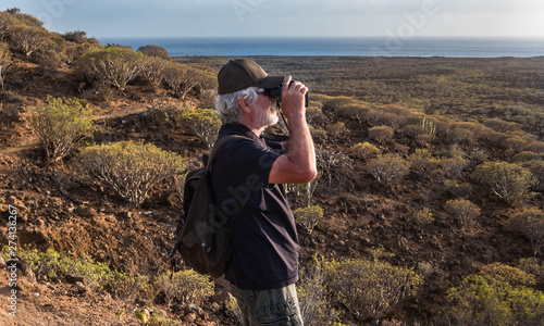 Senior 70 years old man hiking in the mountain and stops to look the landscape with a binoculars. Happy excursion outdoor. Volcanic landscape and blue sky