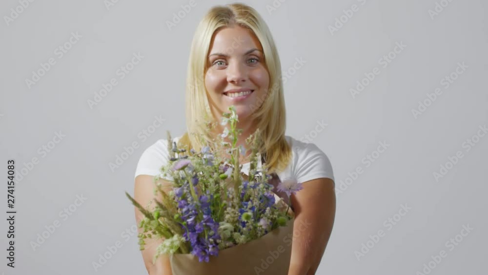 Chest-up portrait shot of delighted 20-something blonde florist posing against light-grey wall and presenting her dainty fresh wildflower and delphinium bouquet, wrapped in brown craft paper Chest-up portrait shot of delighted 20-something blonde florist posing against light-grey wall and presenting her dainty fresh wildflower and delphinium bouquet, wrapped in brown craft paper