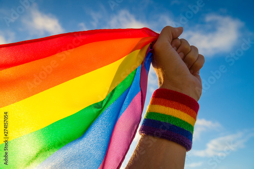 Hand with rainbow wristband waving gay pride flag waving backlit in the wind against a bright blue sky