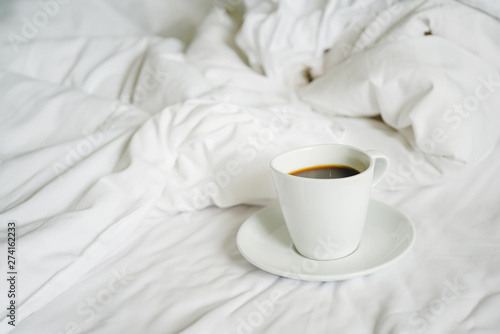 white ceramic cup of hot black coffee on messy bed sheet, in soft focus