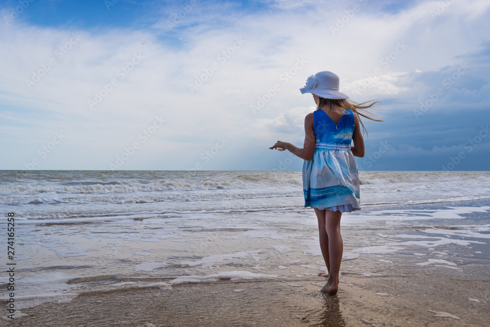 A girl in a beautiful dress and a white hat with a shell in her hands on the shore against the blue sky background