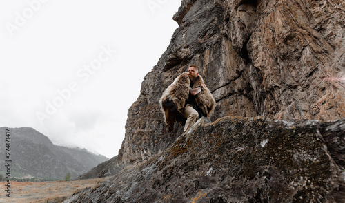 Foto a serious and strict man with a beard and mustache in a black T-shirt and a bear skin stands at a cliff, a mountain