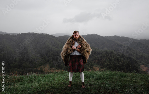 man with mustache beard is standing on top mountain kilt. bear's skin on the back. in a white shirt, socks a tattoo on his arm. on the grass. there is snow.leather bracelet